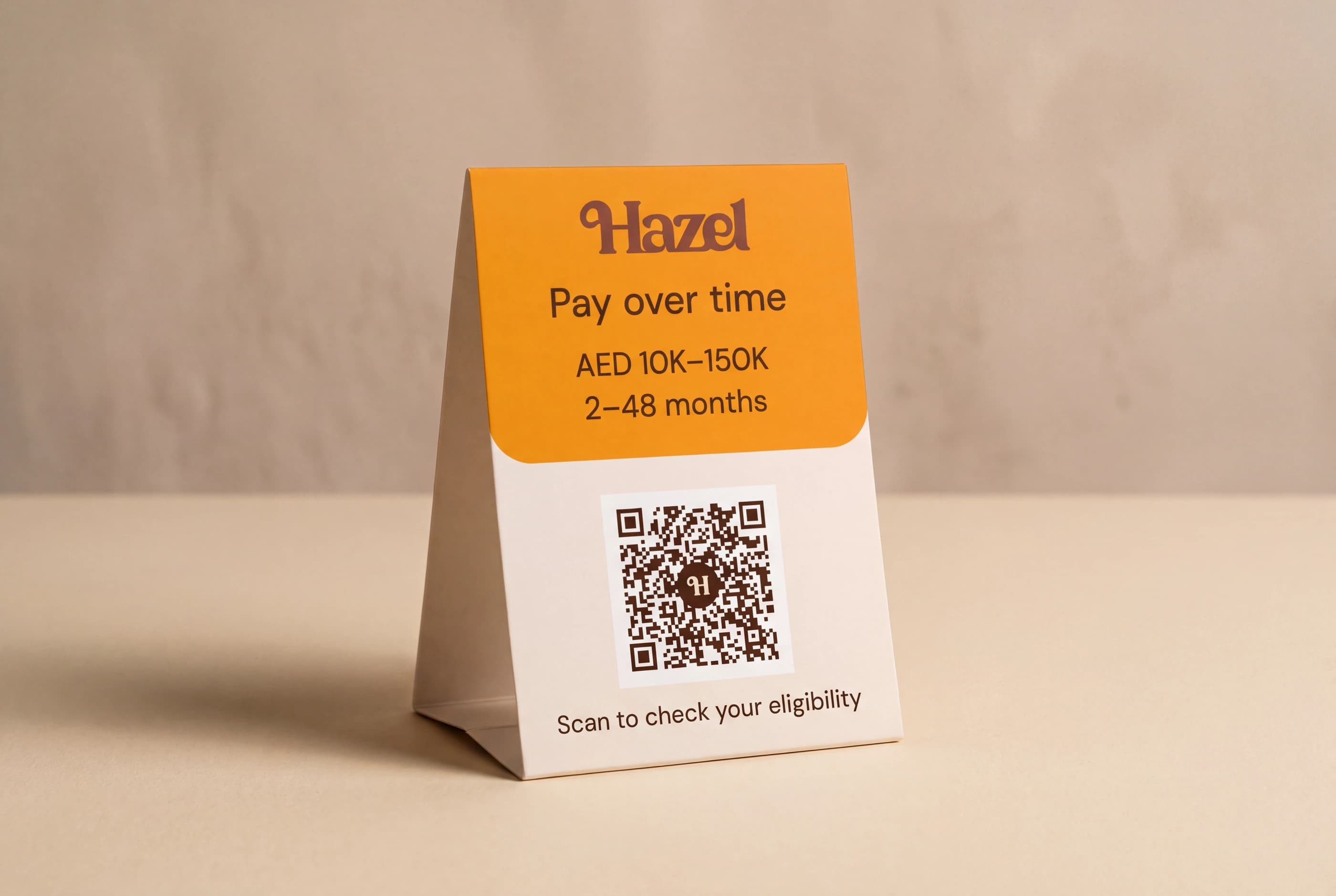 A studio photograph of the Hazel counter card, printed with AED 10K–150K over 2–48 months above a QR code and the line “Scan to check your eligibility”