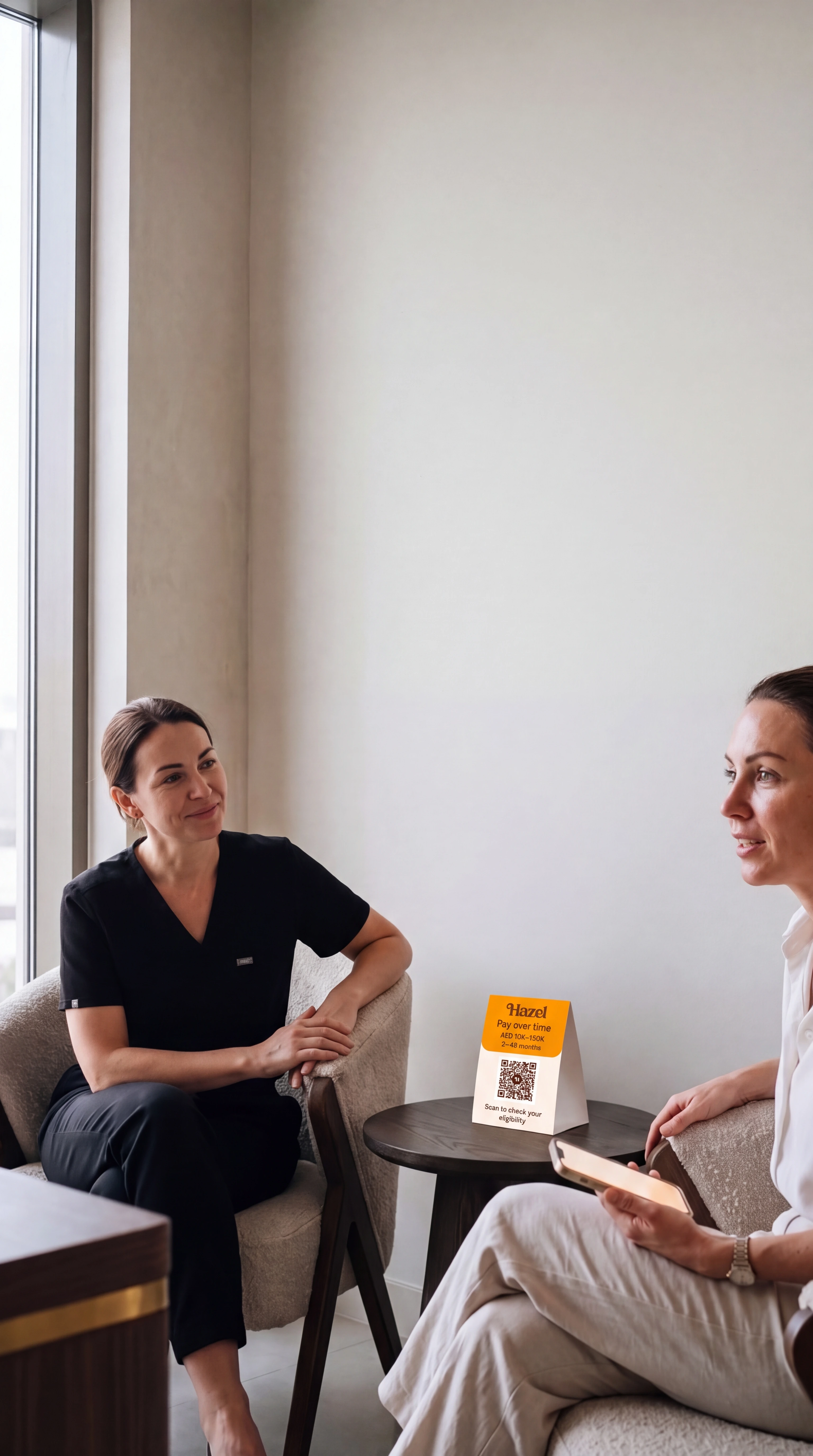 Two people talk in a calm consultation room, one holding a phone, with a Hazel card on the low table.