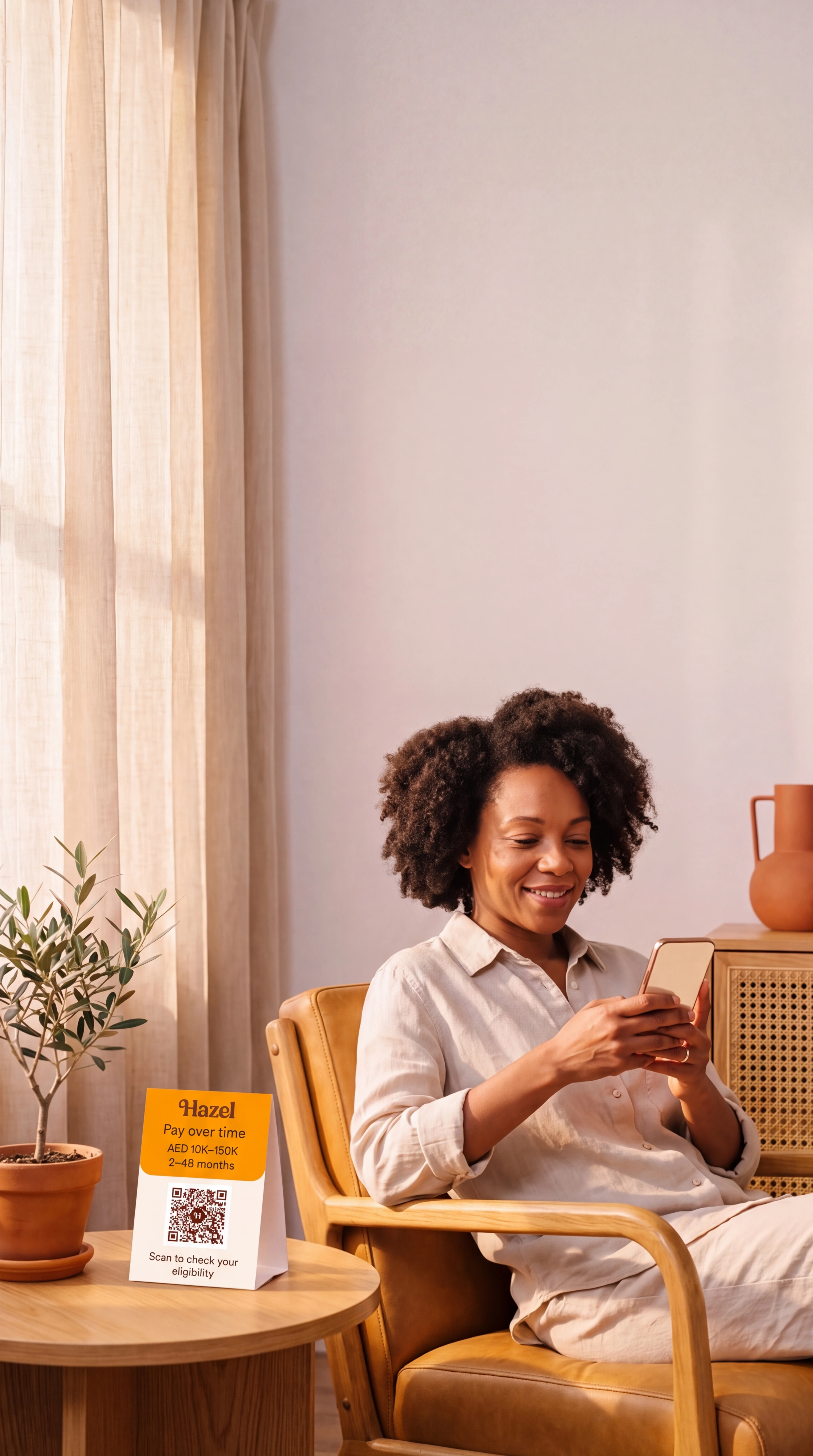 A patient sits in an armchair by a sunlit window, looking at a phone, with a Hazel card on the side table.