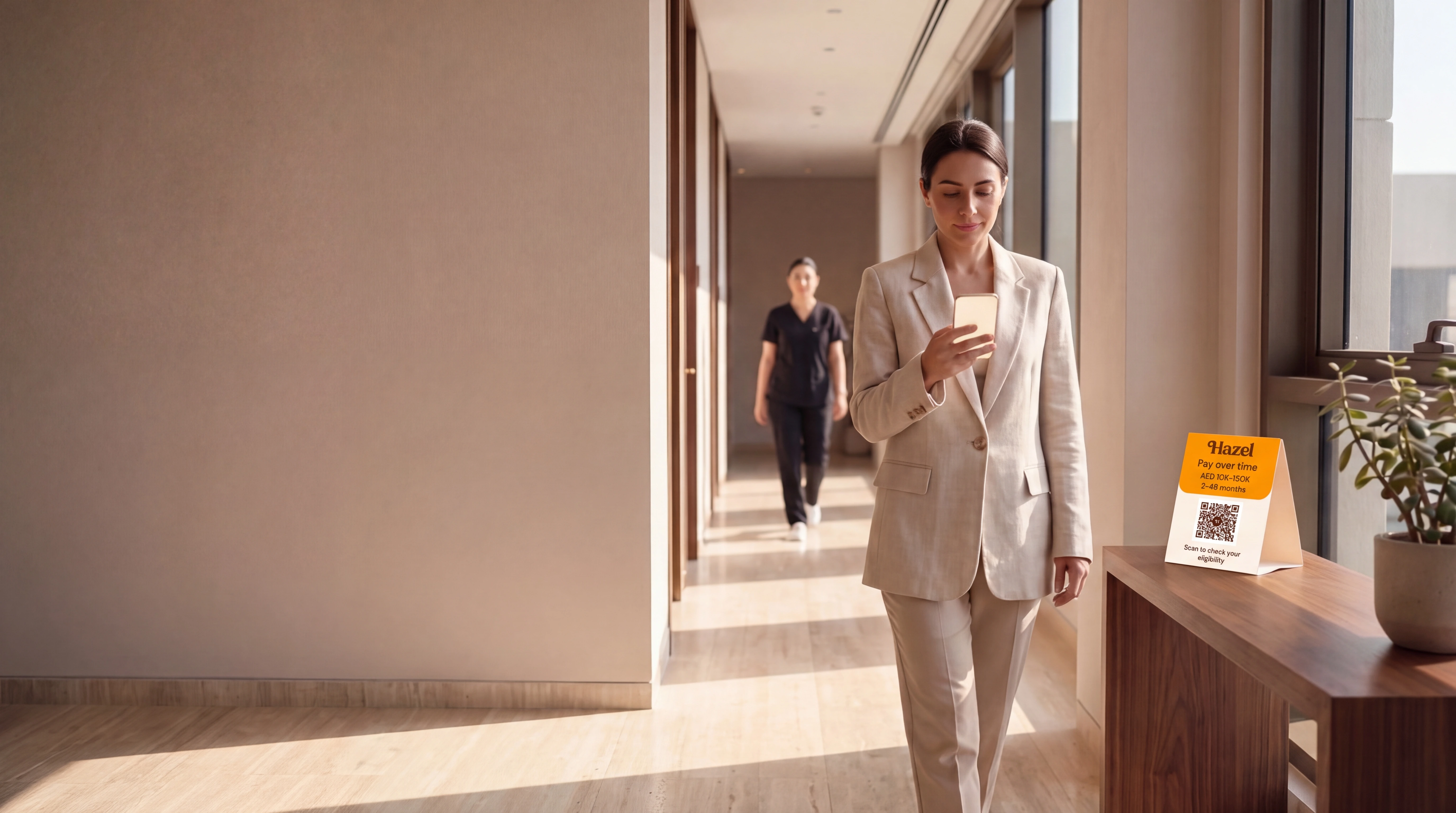 A patient checks a phone in a calm clinic corridor, with a Hazel card on the console alongside.